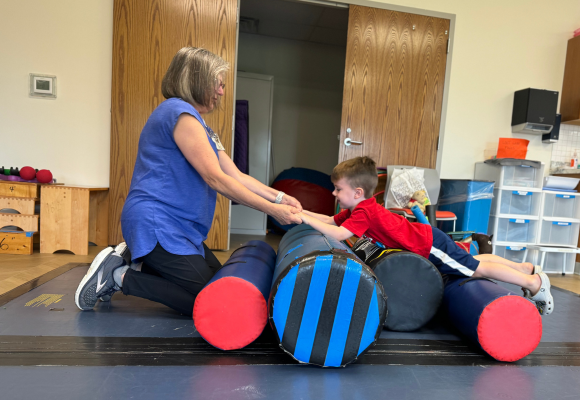 Grayson laying on his stomach on top of four cylindrical rollers on their side, all of different widths, with occupational therapist Bernie holds his hands to help pull him over the rollers. 