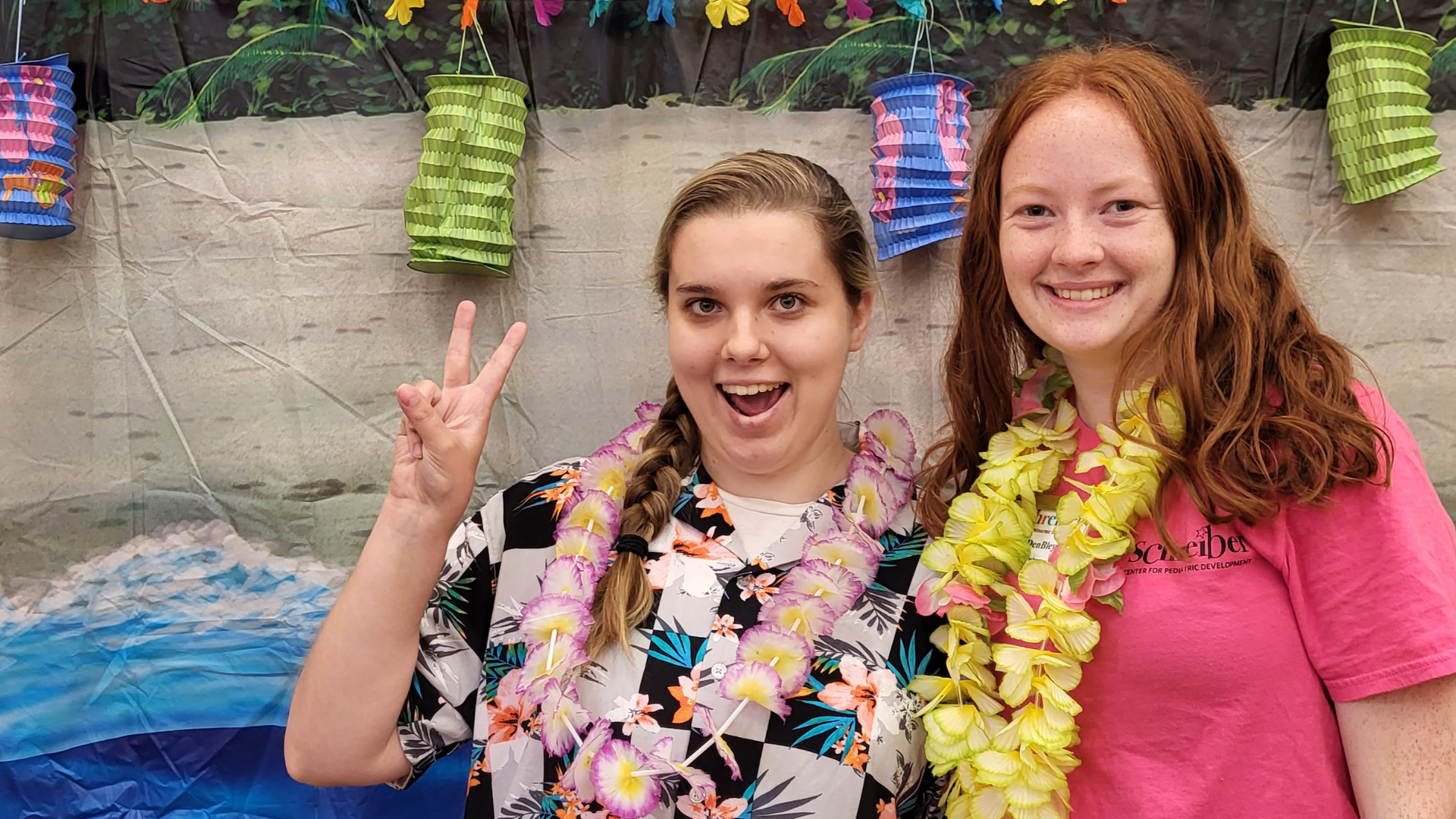 Two tweens smiling and one is holding a peace sign up, dressed in tropical outfits.