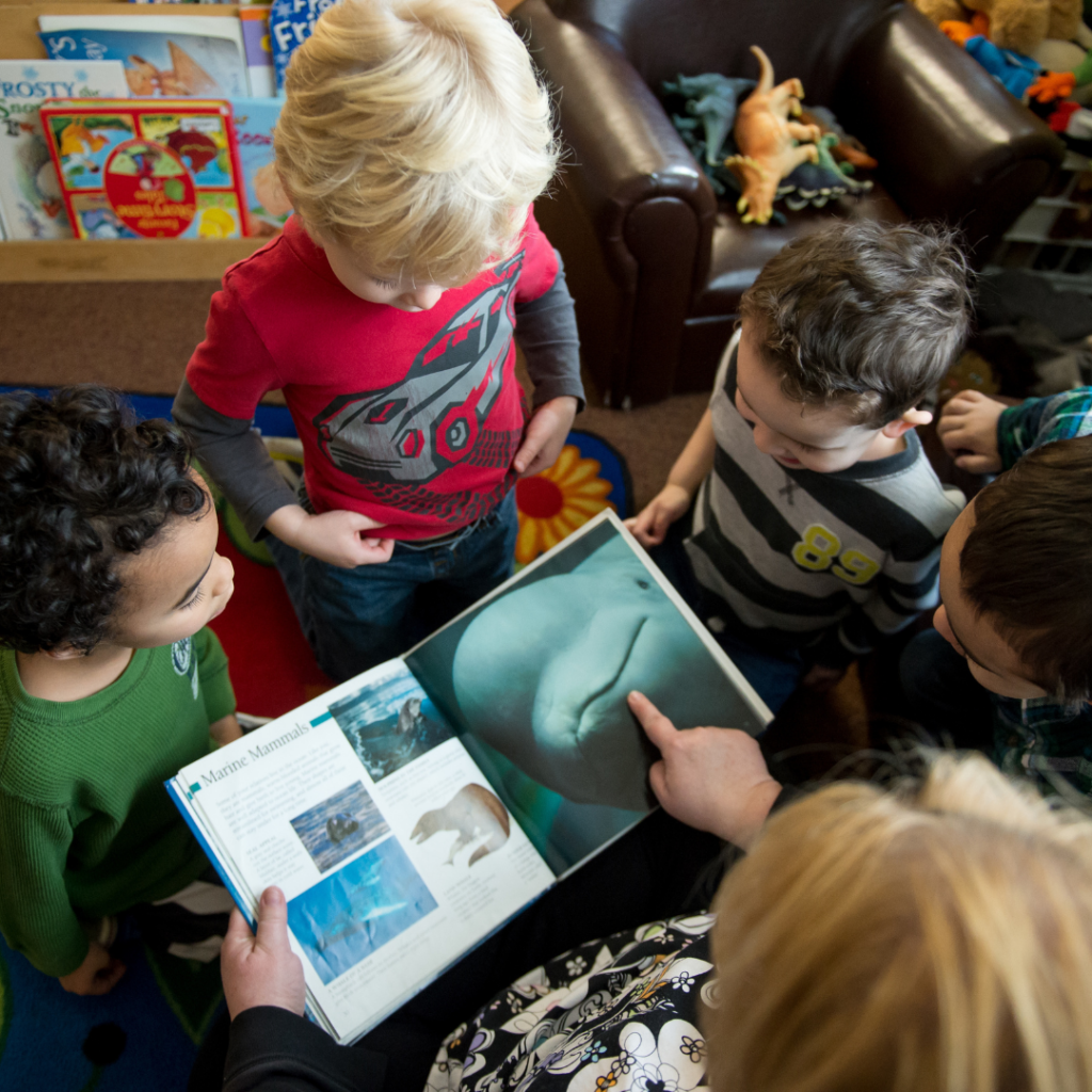 A group of preschool age children surrounding a book being held by an adult.