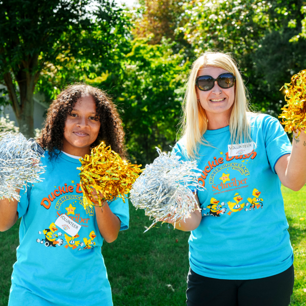 Two people dressed in duckie race t-shirts holding up silver and gold pompoms.