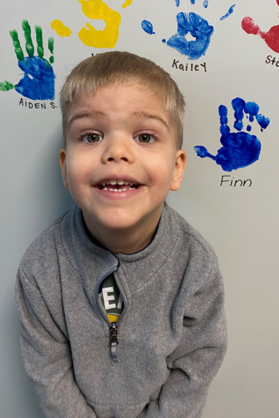 Finn smiling and standing in front of his blue graduation handprint on a wall.