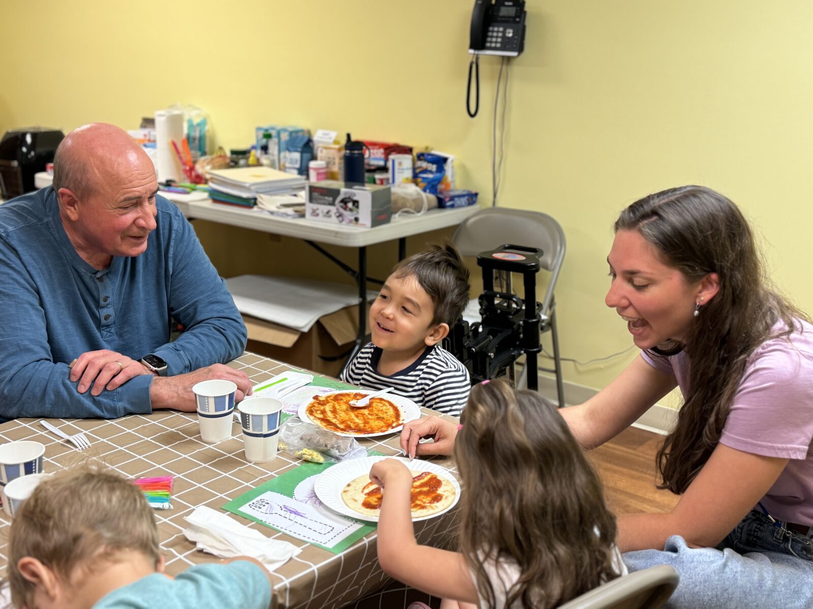 Adults and children smiling around a table spreading sauce on pizzas.