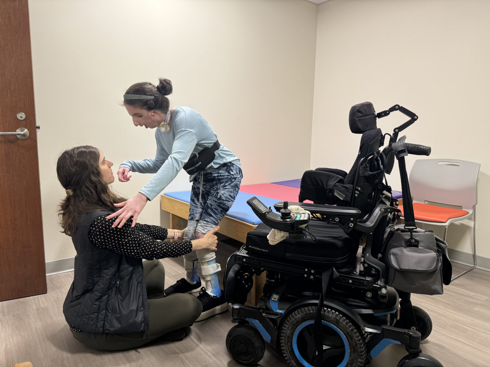 A therapist assisting a child perform physical therapy exercise with a wheelchair nearby.