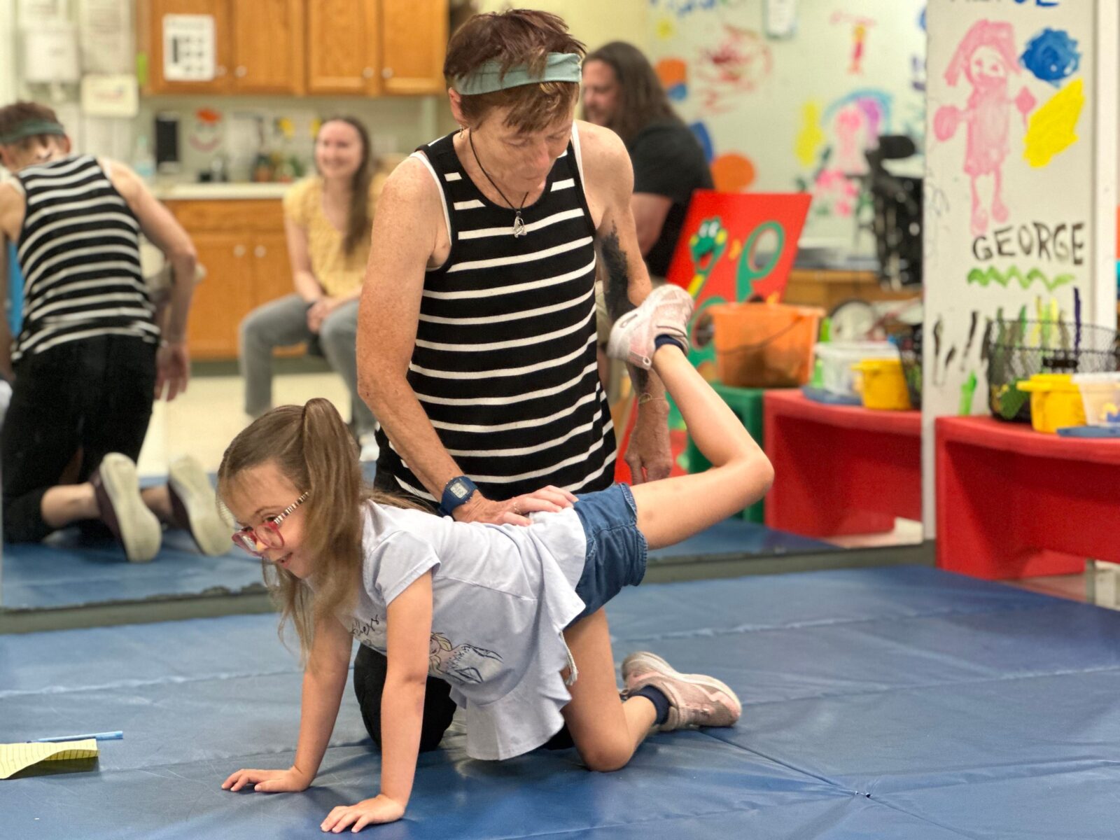 A therapist guiding a child through a physical therapy exercise where they kick their leg in the air on a blue exercise mat.