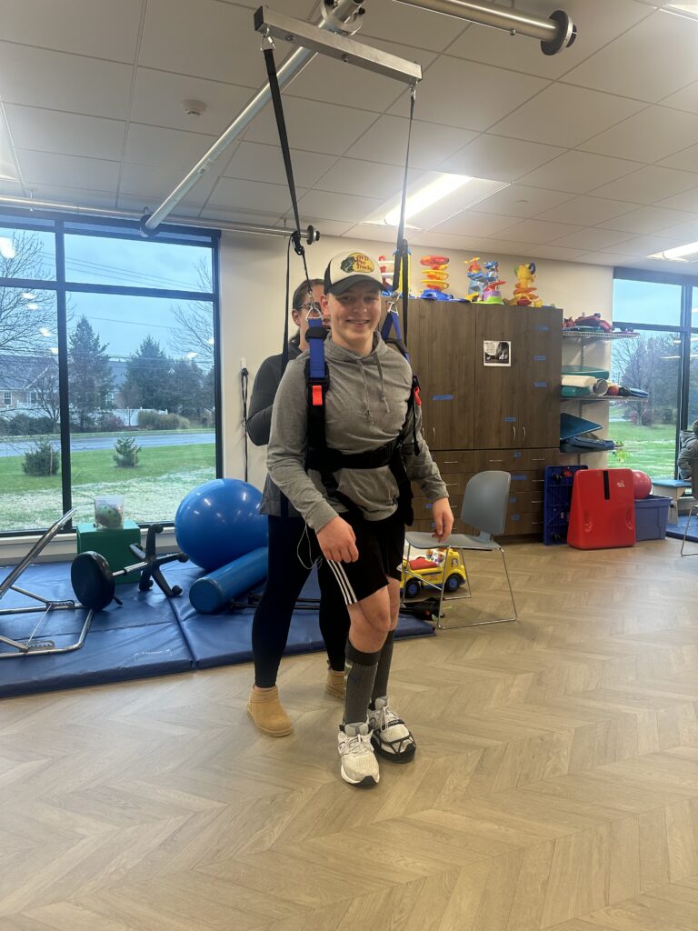 A child smiling while using an overhead track harness with a therapists assistance.