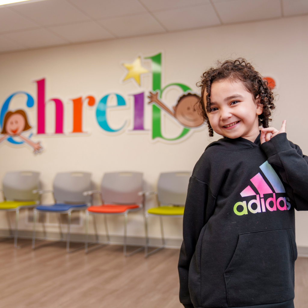 Schreiber student in black adidas sweatshirt smiling in front of Schreiber logo wall