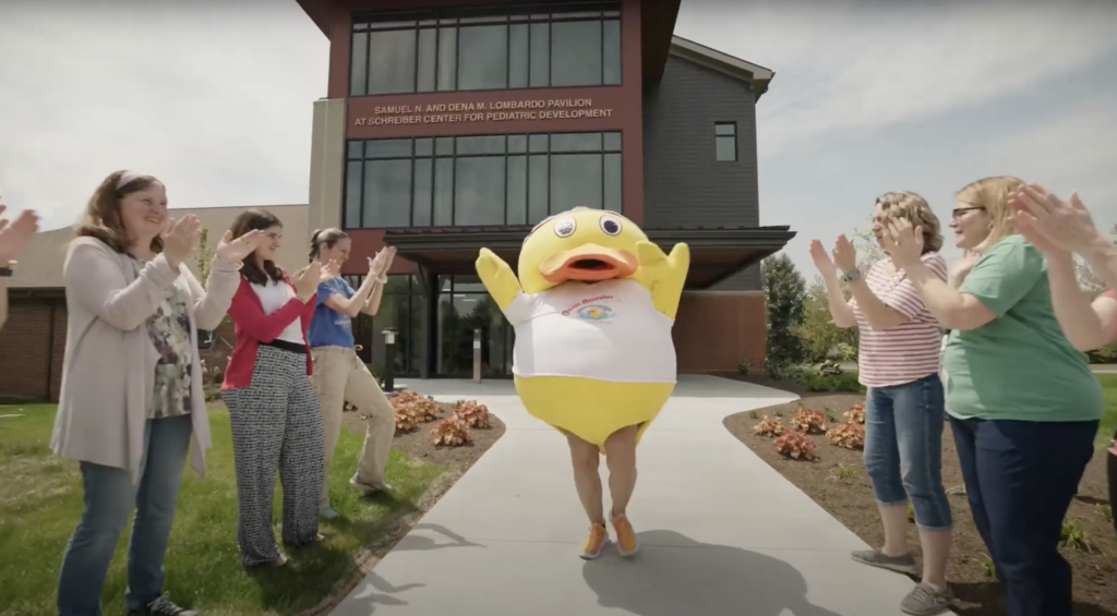 duck mascot running down sidewalk in front of the Schreiber building giving high-fives