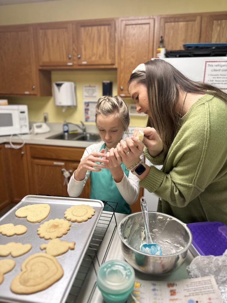 A kiddo and adult using pastry bags to add frosting to cookies on a cookie sheet.