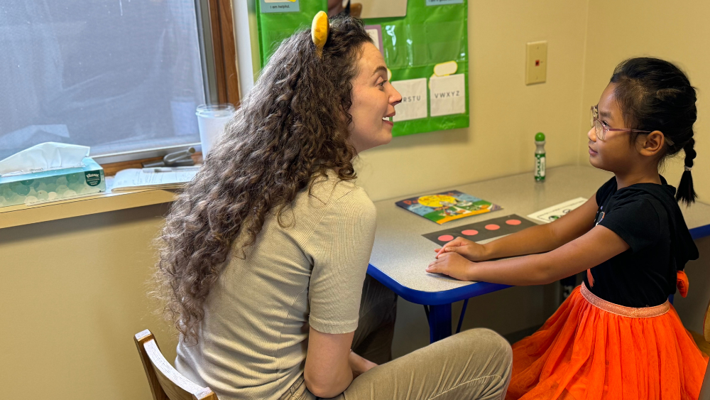 A child with glasses and a braid working at a table with her speech therapist.