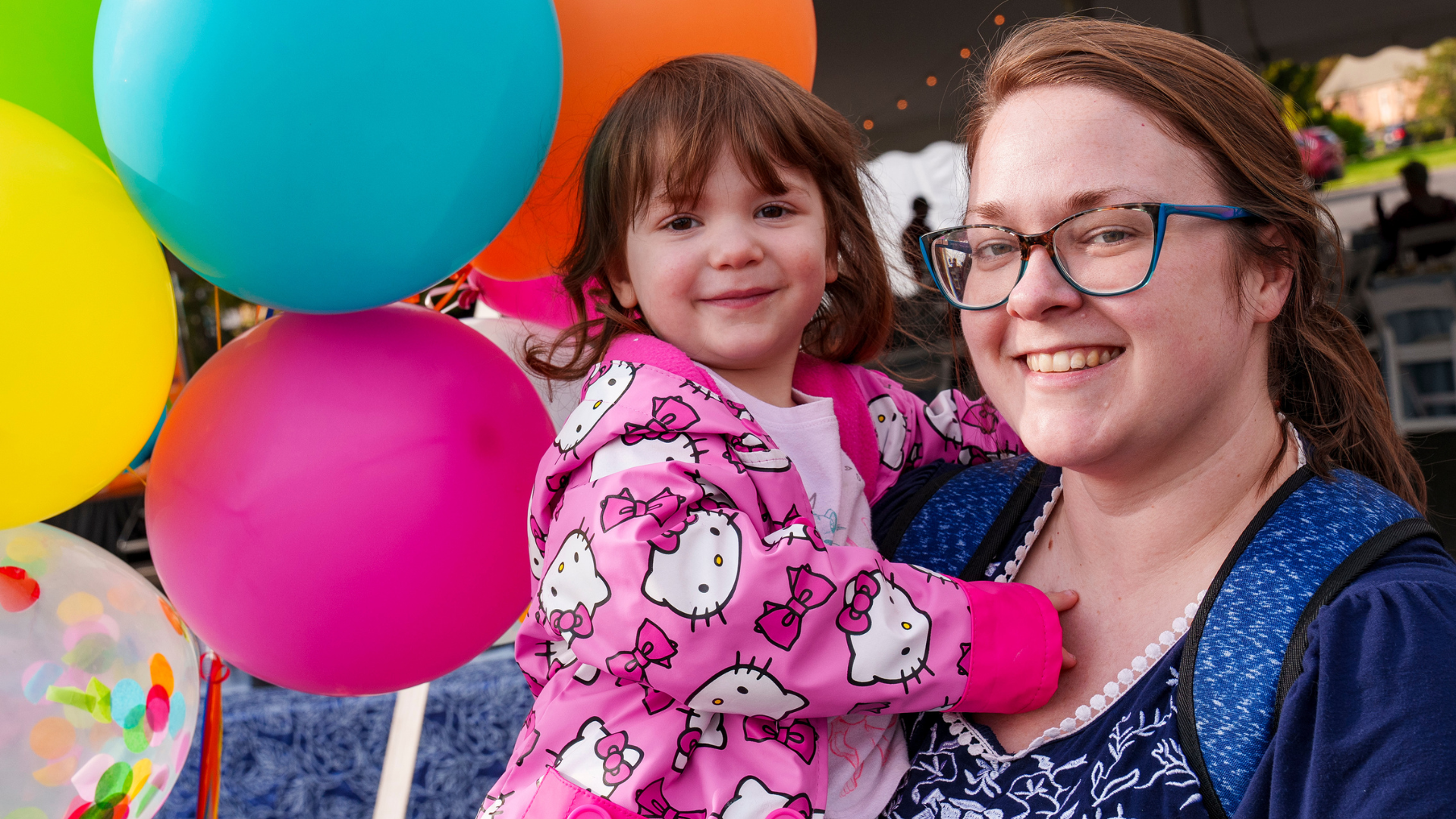 A child being held by an adult, both smiling, in front of bright yellow, blue, and pink balloons.